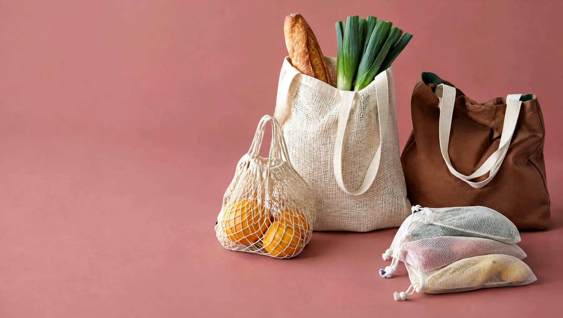 Four cotton bags on a dusty-rose background: a market tote, a string net, a shoulder tote and produce bags