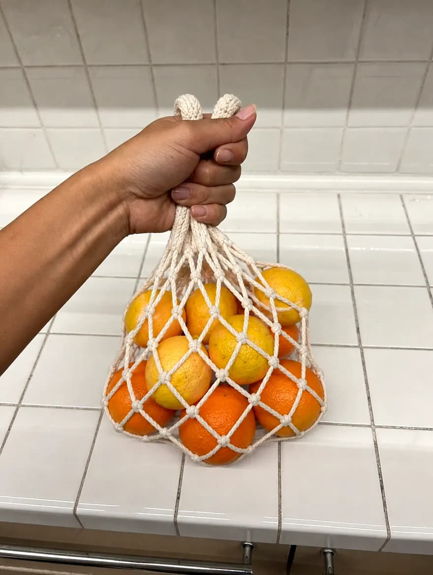 A hand lifting a cream cotton string bag full of citrus off a tiled counter